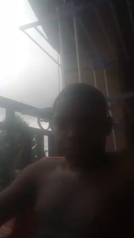 A shirtless person with short, dark hair is seated in an orange chair. Behind the person is a white clothes drying rack. A railing is also visible. The scene is brightly lit.