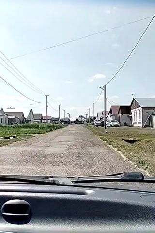 The video shows a view from inside a car, looking forward through the windshield. The car is driving down a paved road that appears to be in poor condition with visible cracks and potholes. On both sides of the road, there are houses, mostly single-story with peaked roofs, and some with fences. There are utility poles with wires running along the street. A few cars are parked on the side of the road and some are visible driving in the distance. The sky is blue with a few scattered clouds, suggesting it is daytime and likely clear weather. The overall impression is of a rural or suburban street.