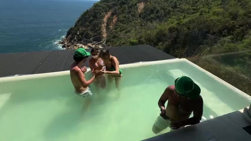 Four individuals, two males and two females, are seen relaxing in a swimming pool on a sunny day in Puerto Marqués, Mexico. The pool, featuring light green water and an infinity edge, overlooks the vast blue ocean and a lush, green-covered cliffside.

The two males are positioned on the left and right sides of the pool, both wearing green baseball caps. The cap on the male to the left has partially visible text "MAKE MEXIC...", while the cap on the male to the right clearly reads "MAKE MEXICO GREAT AGAIN". Both males are holding smartphones, seemingly capturing moments or interacting with their devices.

The two females are situated in the center of the pool. They are wearing bikinis and holding small drinks or objects. Their hair is slightly ruffled, suggesting a gentle breeze. The group appears to be in good spirits, enjoying their time together in the scenic outdoor setting. The sound of wind is audible, adding to the ambiance of the coastal location.