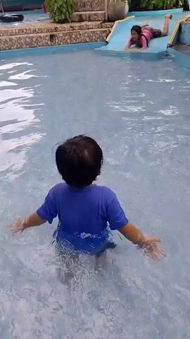 A toddler in a blue shirt and blue shorts stands in a swimming pool. The toddler faces away from the camera and towards a blue water slide where a young girl in a pink bathing suit is on the slide. The water is clear and blue. The pool area is tiled with rocks.