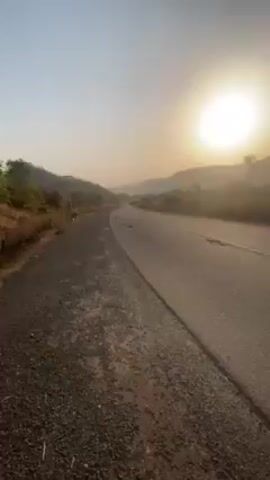 The video begins with a shot of a road leading away from the camera toward mountains on the horizon, with the sun high in the sky above them. The camera is positioned on the side of the road, and moves forward slightly. A man wearing a light blue outfit walks past the camera on the road. A tire and some kind of leafy vegetation are lying on the road. The man is walking away from the camera.