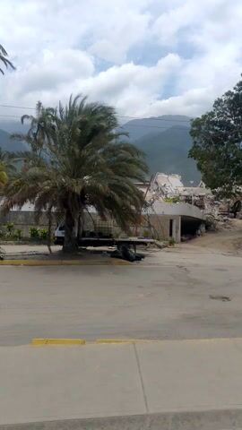 The video displays a roadside scene with a variety of elements. A large palm tree dominates the foreground, its fronds appearing dry and possibly wind-battered. In the background, mountains rise against a sky filled with clouds. There are also other palm trees visible. A damaged or unfinished building is partially obscured by vegetation. A white truck is parked on the side of the road. A man and two women are engaged in physical activity, possibly exercise, on the paved area. They appear to be a short distance from the tree. The road has yellow lines painted on its edge, indicating a parking or traffic demarcation. The overall environment suggests a tropical or sub-tropical location with signs of construction or decay.
