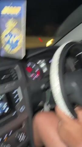 Inside a dark car, the driver's hands are on a rhinestone-studded steering wheel while loud music plays. An air freshener hangs from the rearview mirror. The camera briefly focuses on the two male passengers in the back seat, then swings to a man in the front seat wearing a white shirt, before returning to the car's dashboard.