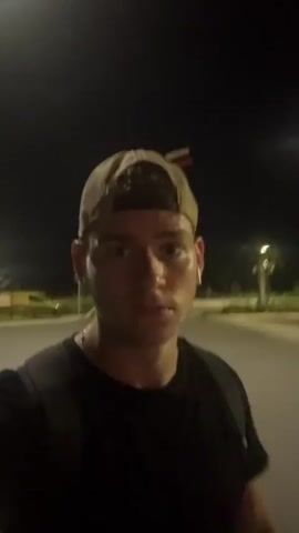 The media file is a selfie video featuring a young man wearing a baseball cap and a black t-shirt. He is standing outdoors at night, possibly in a parking lot or open area, given the visible streetlights and paved ground. The man is looking directly at the camera with a neutral expression. He has a backpack on. In the background, there are more streetlights and some trees, suggesting an urban or suburban environment. The sky is dark, with a visible moon. The overall impression is that the man is taking a casual video, perhaps while out for a walk or on his way somewhere. The lighting is dim, indicative of nighttime. There is no visible text in the media file. The location context provided is Panama City Beach, United States of America, which aligns with the possibility of an outdoor setting at night.