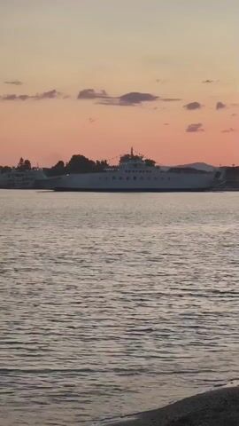 A large white ferry travels across a body of water. The sky above the ferry is a gradient of orange and pink hues, with a few scattered clouds. The water in the foreground is rippled and appears to be reflecting the colors of the sky. The shoreline is visible in the bottom right corner of the frame, composed of small stones. Trees and buildings are faintly visible on the distant shore behind the ferry.