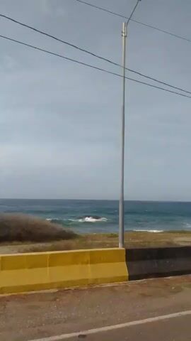 A video shows a view of the ocean from a moving vehicle. The ocean is blue with white waves crashing against a rocky shore. There is a yellow and black barrier along the side of the road. The sky is overcast.