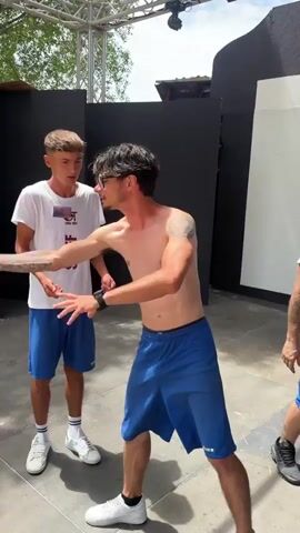 Two young men are standing in an outdoor area, possibly backstage or a service area, given the metal truss structure overhead and dark panels in the background. The man on the left, wearing a white t-shirt with "STAFF" printed on it and blue shorts, is facing the man on the right. The man on the right, shirtless and wearing blue shorts, is facing the man on the left, gesturing with his hands. Both men are wearing white sneakers. The ground appears to be concrete or pavement. The lighting suggests it is daytime. The overall impression is one of casual interaction or rehearsal.