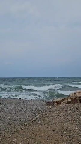 The video shows a rocky beach with the ocean in the background. The waves are crashing on the shore, and the sky is cloudy. There are several large rocks on the beach, some of which are partially submerged in the water. The water is a deep blue-green color, and the waves are white-capped. The overall mood of the video is peaceful and serene.