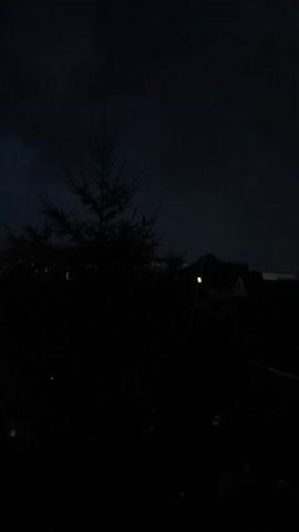 This is a short video taken at night. The scene is very dark and features a silhouette of a spruce tree in the foreground, with what appears to be houses in the background. There is a single lit window visible in one of the houses. The sky is dark and cloudy. The overall mood is dimly lit and somewhat ominous.
