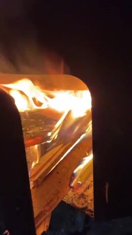 A close-up shot of a fire burning in a metal fire pit. The flames are bright orange and yellow, with some wisps of smoke rising upwards. The fire is contained within a black metal structure that has an opening in the front, allowing the flames to be seen. Several pieces of wood are visible in the fire, and they are partially burned. The background is dark, suggesting that it is nighttime or a dimly lit area. The camera is positioned at an angle looking into the fire pit.