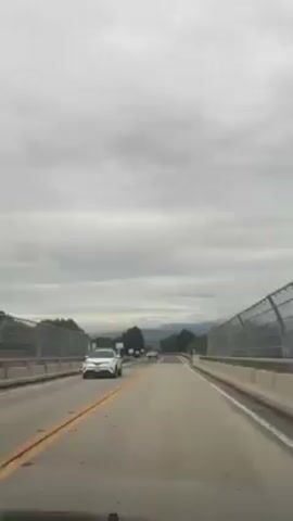 The video shows a driver's perspective of driving on a two-lane highway with a yellow dividing line. The road is elevated on a bridge structure with metal safety fencing. Several cars are driving on the road ahead. The surrounding landscape consists of trees and rolling hills under an overcast sky. The interior of the car is visible.