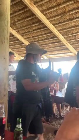A man wearing a white t-shirt with a skull and crossbones design is standing in a beachfront bar. He has money pinned to his shirt. He is gesturing with his hands. Two other people are visible in the background, one wearing a striped shirt and the other in a black shirt. The bar has a thatched roof.