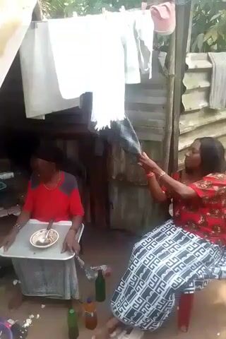 The video shows a group of people gathered in what appears to be an outdoor area near a structure with a corrugated metal roof.  Clothes are hung on a clothesline. A man in a red shirt sits at a small table with a plate and spoon.  Another man in a red tank top and patterned pants crouches near a bucket.  Several women are also present, one holding a phone. People are milling around and talking.
