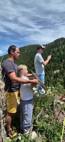 A group of people and a dog are on a mountain trail. A man is standing to the left, holding a young girl's arm. She is holding a flat, rectangular object, possibly a piece of equipment, and looking towards the right. Behind them, a man in a cap is taking a picture with his phone. A dog is sitting on the edge of the trail to the right, on a leash. The background is a steep, forested mountain with a cloudy blue sky. The foreground consists of a dirt path with wildflowers.
