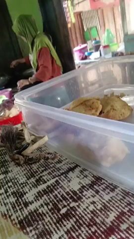 A close-up shot shows two fried pastries in a clear plastic container. In the background, a person wearing a green headscarf and an orange shirt is visible. Further in the background, there are shelves with various items. The scene appears to be an indoor setting, possibly a food stall or a home. The camera then pans to show a person's hand holding a pitcher of liquid. The pitcher is partially obscured by the pitcher's spout. The video then shifts to a wider shot of a table with a patterned tablecloth. There are several people in the background, some of whom are sitting. A woman in a patterned shirt is in the foreground, looking to the side. A blue bowl with some green leafy vegetables and a spoon is on the table in front of her. A glass with a yellow straw and a bottle with a teal cap and a metal spoon are also visible. The lighting suggests daytime.