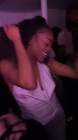 The video shows a group of young adults partying in what appears to be a room with purple and blue lighting. People are dancing and singing along to music, and many are holding red cups. The camera moves around, capturing different individuals in the crowd, many of whom are smiling. The environment suggests a lively and social atmosphere.
