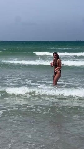 The video shows a woman in a black bikini walking into the ocean. The ocean has small waves breaking on the shore. The sky is overcast. There are a couple of ships in the distance. The camera zooms in on the waves and then back to the woman as she continues into the water.