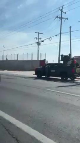 The video shows a convoy of military vehicles on a highway next to a high fence. The vehicles are painted in camouflage and have soldiers standing on top. There is a road barrier blocking the road. The sky is blue with some clouds and power lines run parallel to the road. The video is taken from a moving vehicle on the opposite side of the road.