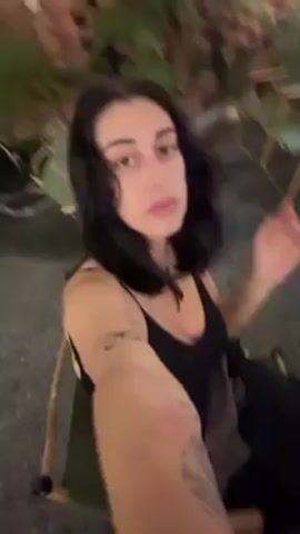 A person with dark hair is taking a selfie outdoors. They are wearing a black tank top and sitting on a small, folding camp chair. The background is a paved surface with other tables and chairs visible. They run their hand through their hair, making adjustments. The person has tattoos on their arm that is extended towards the camera.