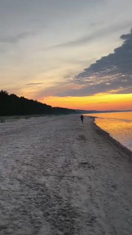 The video captures a serene beach scene at Apšuciems, Latvia, during a sunset. The sky is a canvas of vibrant colors, transitioning from a fiery orange and yellow at the horizon to soft pinks and purples higher up, with scattered clouds adding depth to the spectacle. The calm sea mirrors the sky's hues, creating a breathtaking reflection.

A lone figure is seen walking along the sandy shore, adding a sense of solitary contemplation to the vast landscape. The beach itself is wide and sandy, with a gentle slope leading into the tranquil water. The distant treeline suggests a forested area bordering the coast.

The time of day is clearly sunset, indicated by the warm, golden light and the rich colors in the sky and on the water. The weather appears to be calm, with minimal wind suggested by the smooth surface of the sea and the gentle ripples. The overall mood is peaceful and reflective, evoked by the natural beauty of the setting and the solitary presence of the individual. There is no visible text in the video.