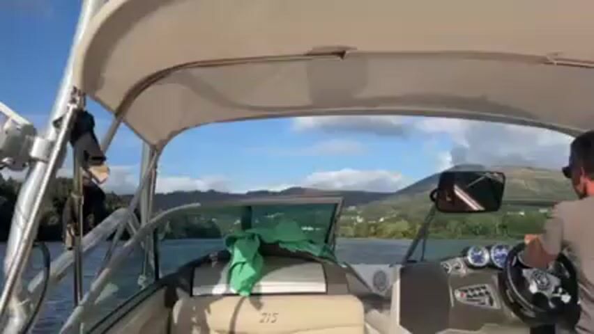 The video shows a person driving a motorboat across a body of water. The person is wearing sunglasses and a grey t-shirt. The boat has a canopy, windshield, and dashboard with gauges and buttons. There's a green towel on the passenger seat. The background features green trees, land, and a partially cloudy blue sky. The boat is moving at a moderate speed, creating waves in the water.