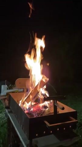 A large fire is burning in a rectangular metal brazier at night. Sparks fly up from the fire. The flames are bright yellow and orange, with some red embers visible at the base. The brazier sits on a table outdoors, with some green foliage visible at the bottom of the frame and a dark background suggesting a nighttime setting.