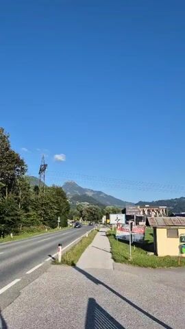 The video shows a roadside scene on a clear, sunny day. The camera is positioned at an intersection, looking down a paved road that curves to the left. To the right of the road is a sidewalk, followed by a small yellow building with a green trash receptacle. Further to the right, there are commercial buildings with signs, and in the distance, a large, tree-covered mountain dominates the horizon. A traffic sign prohibiting motorcycles and mopeds is visible on the right side of the road. There are a few cars driving on the road in the distance, and a single cloud floats in the bright blue sky. The overall impression is of a rural or semi-rural setting.