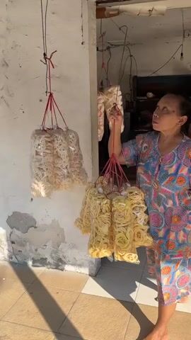 A middle-aged woman with dark hair tied back is standing in front of a white, weathered wall. She is wearing a colorful, patterned dress and matching shorts. She is holding a bundle of clear plastic bags, each filled with yellow, ring-shaped snacks, likely crackers or chips. The bags are tied together with red string and suspended from a black wire attached to an overhang. The woman is looking up and to her left, with her right hand reaching towards the hanging bags. Sunlight casts shadows on the tiled floor in front of her. The environment appears to be an outdoor or semi-outdoor space, possibly a shop or a home.
