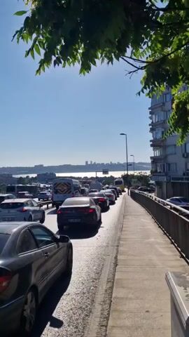 A time-lapse video shows a sunny day with a clear blue sky and some foliage in the foreground. The camera pans across a busy road filled with traffic, which appears to be moving slowly. Cars, vans, and a bus are visible in the lanes, with some vehicles partially obscuring others. A sidewalk runs alongside the road, with a metal railing. In the distance, across a body of water, a city skyline with buildings and bridges can be seen. The sun is casting long shadows, indicating it is likely late afternoon or early evening. The overall impression is of a typical urban traffic scene on a bright day.
