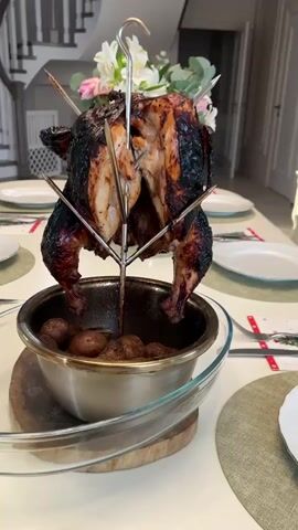 A full, roasted chicken with dark, glistening skin is presented on a metal stand placed atop a bowl of small, round potatoes. The chicken is secured to the stand with skewers, and a floral arrangement adorns the top. The setup is on a white dining table, surrounded by place settings with white plates and silverware. A staircase is visible in the background, contributing to the homey, domestic setting. The camera subtly zooms out, showcasing more of the room and the table setting.