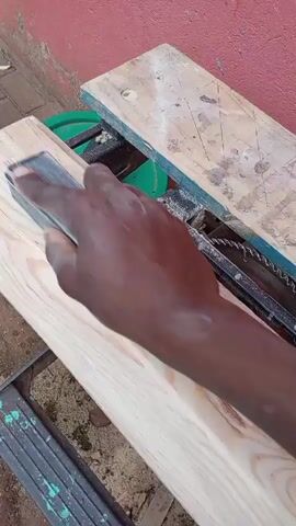 A close-up shot shows a person's hand holding a sanding block and sanding a wooden plank. The wooden plank is positioned on a table saw. The saw blade is visible beneath the plank. The background consists of a reddish wall. The video is filmed outdoors, with some debris on the ground.
