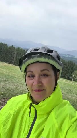A woman wearing a neon yellow jacket and a grey bicycle helmet smiles at the camera. She is outdoors, with a green, grassy hill behind her, a dense forest in the middle ground, and mountains in the background under a cloudy sky.