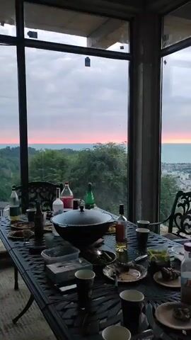 The video shows an indoor gathering with people sitting around a table with drinks and food. The scene shifts to show a different angle of the room, revealing more of the people and the surroundings. The gathering appears to be casual, with attendees enjoying themselves. The room has large windows, offering a view of the outside, including distant hills and what appears to be a sunset or sunrise with a pink sky.