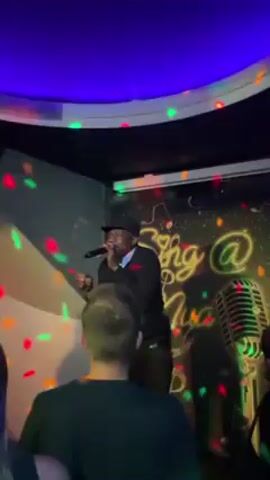 The video captures a lively scene inside a karaoke bar. A man in a black suit and hat is passionately singing into a microphone on a small stage. Red, green, and white disco lights dance around him and the audience, which is gathered closely in front of the stage. Behind the singer, a banner reads "Sing @ Nite Music" with a gold vintage microphone graphic. The camera pans around to show the crowd, the bar area, and the lighting setup, revealing a crowded room with people chatting, drinking, and enjoying the performance. The bar is well-stocked with bottles, and the ceiling is adorned with colored lighting.