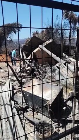The video shows the aftermath of a fire, possibly a bush fire. The first shot is taken through a fence showing the remains of a burned building with charred metal and debris. A person in a blue shirt stands in the background. The next shot is an interior view through the fence, showing damaged metal structures and possibly a staircase. The following shots show the interior of a burned-out building. There is significant fire damage. Three women are visible inside the ruined structure. One woman in a pink cap stands with her hands on her hips, looking around. Another woman embraces a third woman. The overall atmosphere is somber and suggests loss and destruction due to the fire.