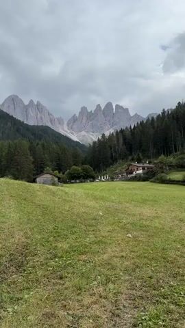 The video shows a panoramic view of a green field in front of a mountain range. The mountains are rocky and jagged, with a few patches of snow. A dense forest of pine trees covers the slopes of the mountains. In the middle ground, there are a few buildings, including a house with a red roof and some smaller structures that appear to be sheds or barns. There are also some parked cars visible near one of the buildings. The sky is cloudy and overcast. The camera is static for the entire duration of the video.