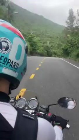 A person wearing a turquoise, white, and red helmet labeled "Leopard Racing" rides a motorcycle along a road that winds around a green mountain. The rider wears a white long-sleeved shirt and a black backpack with "Burton" on it. The motorcycle has two round gauges visible above the handlebars. The road is asphalt with yellow lines marking the lanes. The sky is overcast and the mountain is covered in trees. Three other motorcycles are visible in the distance, rounding a bend in the road.