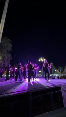 A group of about 16 people stand on a stage, illuminated with purple lighting, at night. The group is facing away from the camera towards an audience, visible in the background.  They are wearing black clothing and white sneakers. The stage appears to be made of dark wood.  A tree and a lamp post are visible to the left of the stage. Palm trees and tents can be seen in the distance behind the crowd. The sky is dark. The stage has metal support beams visible beneath. One person in the group appears to be playing a trumpet. Most of the people have their hands together and are clapping.