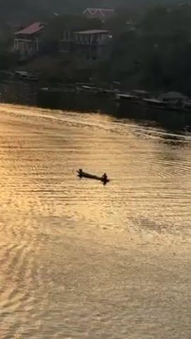 The video shows a person in a narrow boat standing and propelling it with a paddle on a golden river. There are buildings in the background on a hill. The water has many small ripples. Other boats are docked along the edge of the river in the background.