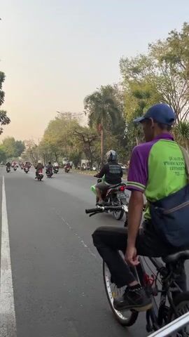A person is riding a bicycle on the right side of a paved road, pedaling with one foot while balancing the other on the pedal. The person is wearing a purple and green shirt, dark pants, and a blue baseball cap. A dark-colored bag is slung across their back. The road has a white line marking the edge of the lane. Several motorcycles are visible in the background, moving in the same direction as the cyclist. There are trees and buildings lining the road, and the sky is overcast. The overall environment appears to be a street in a city or town. The video is taken from a moving vehicle behind the cyclist.