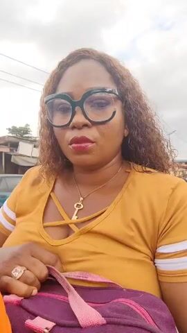 A woman wearing large, dark-green glasses, a yellow top with white stripes on the sleeves, a gold key necklace, and a ring on her left hand is recording a video. She is standing outside in what appears to be a busy street in Nigeria, judging by the yellow-and-black commercial vehicles driving past. She is holding a purple bag. The weather seems overcast.