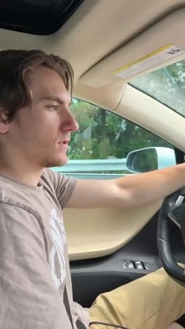 A young Caucasian man with light brown hair is driving a car. He is wearing a light brown t-shirt with a graphic print on the left side and khaki pants. He is gripping the steering wheel with both hands and appears to be speaking angrily to the camera, which is positioned in the car, likely on the dashboard or passenger seat. His mouth is open, and his brow is furrowed. The interior of the car is visible, including a portion of the dashboard and a sunroof. Outside the car, trees and a highway barrier are visible. The timestamp on the video reads 00:00.