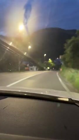 The video shows a view from the inside of a car driving down a road at dusk or early night. The road is paved and has white lane markings. Streetlights are visible along the side of the road, illuminating the surroundings. Trees and buildings are also present. In the distance, a mountain can be seen. A white bus is parked on the right side of the road. The windshield appears to be dirty with streaks. The dashboard of the car is visible in the foreground.