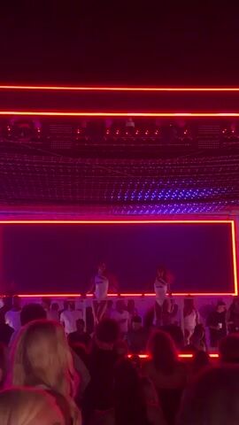 The video shows a crowded event with a stage and a large screen. Two women are on stage, silhouetted against a bright yellow and red background. The crowd is visible in the foreground, with many people's faces blurred by the low light and red lighting. The stage lighting is dynamic, shifting between red, yellow, and purple hues. The overall atmosphere appears to be that of a concert or a club.