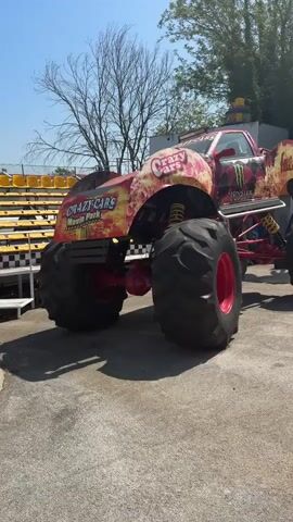 The video shows an area called "Crazy Cars Movie Park" filled with various objects. In the foreground, there's a monster truck with the words "Crazy Cars" on its side, parked on an asphalt surface. Moving towards the center, there's a large yellow and black robot figure resembling "Bumblebee" from the Transformers franchise. Further back, there's an archway with the "Crazy Cars" sign above it, guarded by a red and grey robot figure resembling "Optimus Prime", also from the Transformers franchise. In the background, various vehicles, tires, and other paraphernalia are stacked up, giving the impression of a chaotic junkyard aesthetic. A young girl is seen walking through the archway. The weather appears sunny and clear, and there are trees visible in the background.