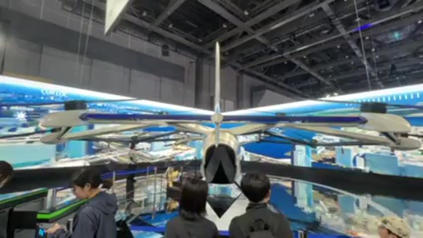 The video shows a large, unmanned aerial vehicle (UAV) with a unique design on display at what appears to be a trade show or exhibition. The UAV has a central fuselage with four horizontal arms extending outwards, each supporting a rotor at the end. Behind the UAV is a large screen displaying a cityscape with weather-related information. Several people are standing in front of the display, observing the UAV. Some are looking directly at the aircraft, while others are looking at the display screens or interacting with other elements in the exhibit. The people vary in age and dress, including adults and a child in a hat. The environment appears to be a large, open space with a high ceiling and visible infrastructure like lighting and ventilation systems.