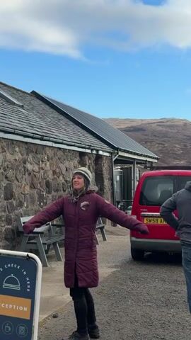 The video shows a sunny day at a gravel campsite near mountains. A woman in a red coat and a patterned hat is standing near a low stone building with solar panels, stretching her arms out to the side. Next to her is a red van with the license plate "SW58 AHL." A man in a gray jacket, jeans, and an orange hat is walking in the same area. In the background, other people are walking around the campsite, and there are various vans, campers, and tents. The sky is blue with white clouds.