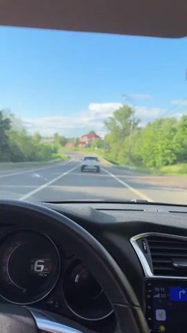 The video shows a driver's perspective while driving a car on a paved road. The dashboard and steering wheel are visible. The driver is following a white SUV in the right lane of a divided road. The sky is blue with light clouds. Green trees line the side of the road. A house with a red roof can be seen in the distance. A second car is visible in the left lane. The navigation system on the car's display indicates that the destination is 3.6 kilometers away. The car's speedometer shows a speed around 60 kilometers per hour and then increases to around 105 kilometers per hour.