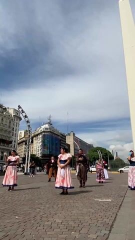 The video shows a group of people in traditional clothing, mostly women in pink dresses with floral patterns, standing on a brick square. A man in brown gaucho attire stands among them. Behind them is the Obelisco de Buenos Aires, a tall white monument. The sky is blue with scattered clouds, and buildings surround the square. Some of the people adjust their clothing and hair. Cars pass by in the background, and a few pedestrians walk nearby.
