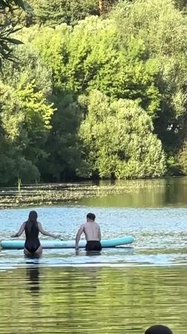 A man and a woman are standing in shallow water next to a paddleboard. The woman is wearing a black swimsuit, and the man is wearing black shorts. The woman is holding onto the paddleboard with her left hand. The man is also holding onto the paddleboard with his right hand. The paddleboard is blue and white. The water is calm and there are lily pads on the surface of the water. In the background, there are trees and foliage. The sky is not visible. The lighting suggests it is daytime.